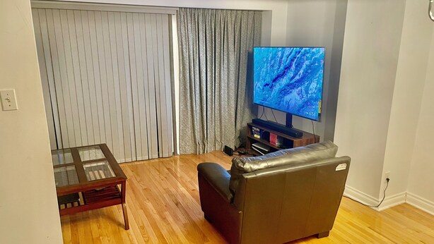 Una sala de estar con un sofá de cuero, una mesa de centro de vidrio, un televisor de pantalla plana y un amplio ventanal con cortinas.