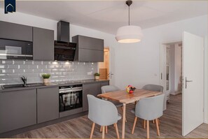 A modern kitchen with grey cabinets, a stainless steel range hood, and a built-in oven. There is a dining table with two grey chairs and a fruit bowl on it.