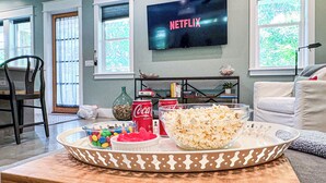 A living room with a large flat-screen TV displaying 'NETFLIX', a coffee table with snacks, a sofa, and a chair.
