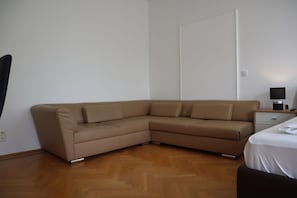 Ein Eckzimmer mit braunem Ledersofa, Holzboden, einem weißen Bett und einem Nachttisch mit Lampe.