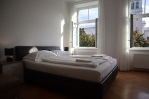 Ein Schlafzimmer mit Bett, Nachttischen und einem Fenster mit Blick auf Gebäude und Bäume.