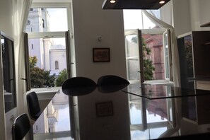 A modern kitchen with a glass countertop, a dining area with chairs, and large windows offering views of buildings and trees.