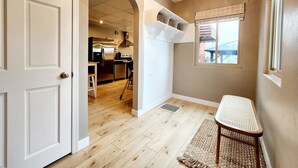 Un pasillo con piso de madera, una puerta blanca, una zona de cocina con refrigerador y estufa, y una ventana con persianas.