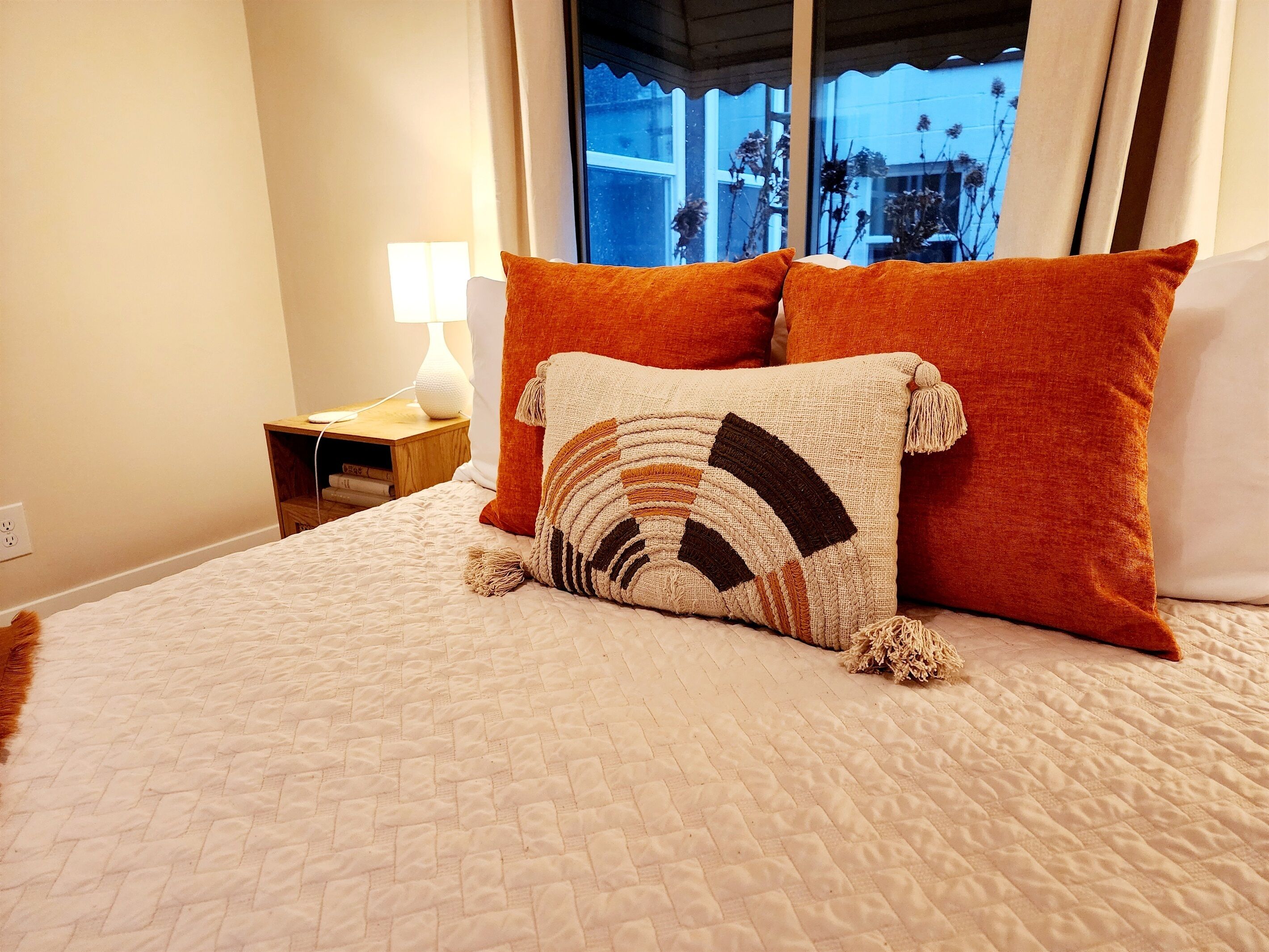 A neatly made bed with a patterned throw pillow, a nightstand with a lamp, and a window with a view of the outside.