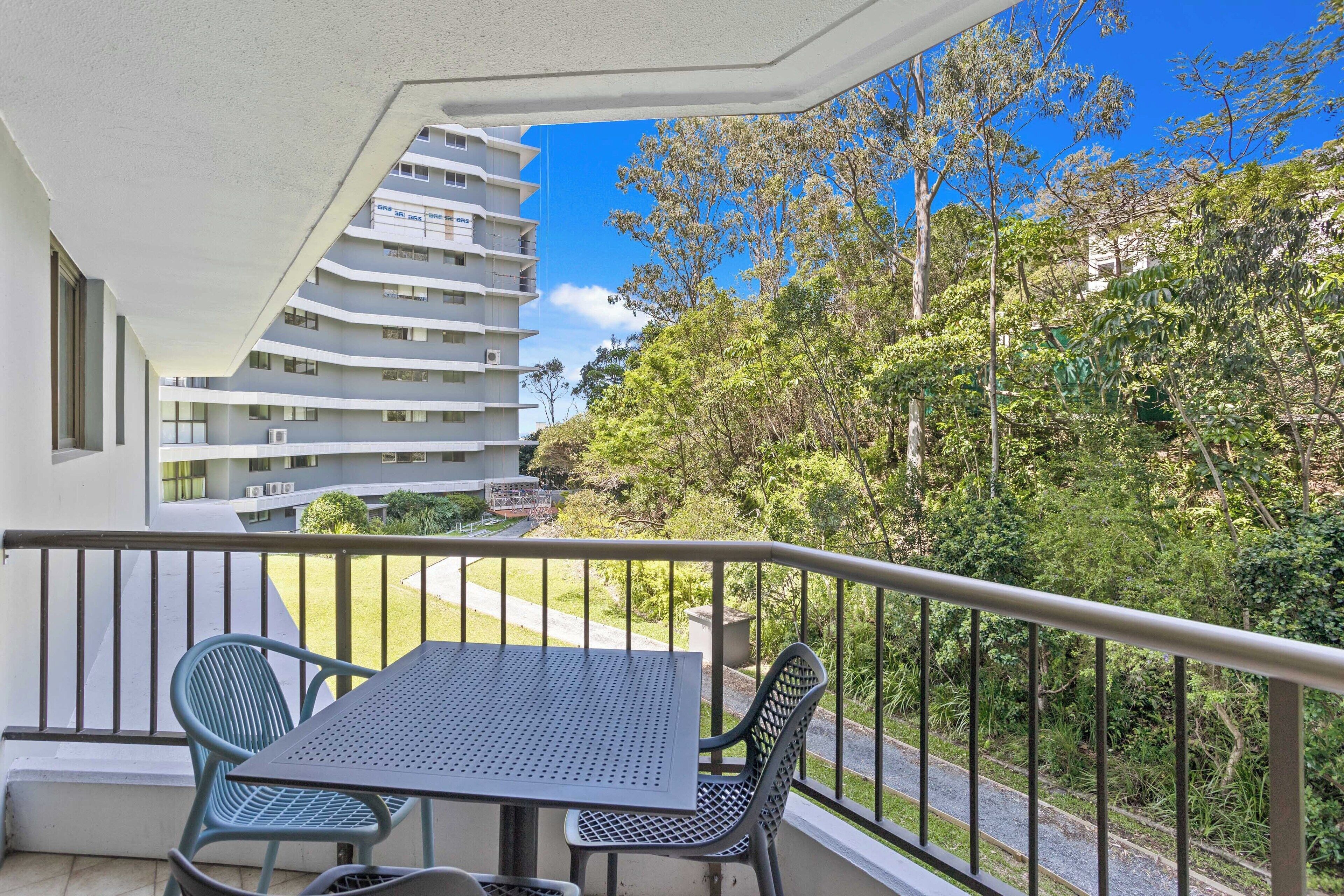 Ein Balkon mit Tisch und Stühlen, der auf eine üppige Grünanlage und ein mehrstöckiges Gebäude blickt.