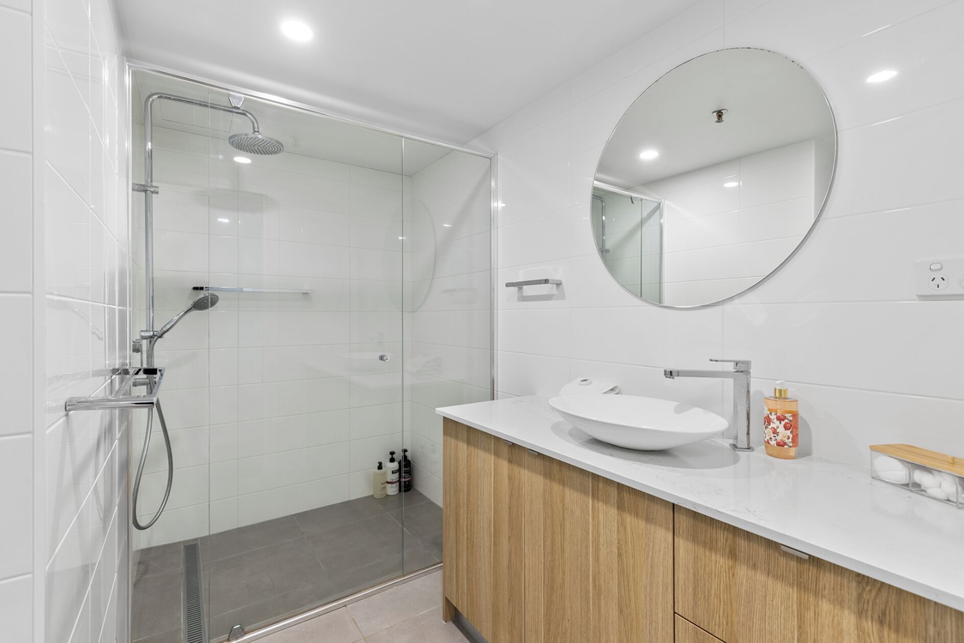 A modern bathroom with a glass-enclosed shower, a round mirror, and a white sink with wooden cabinets.