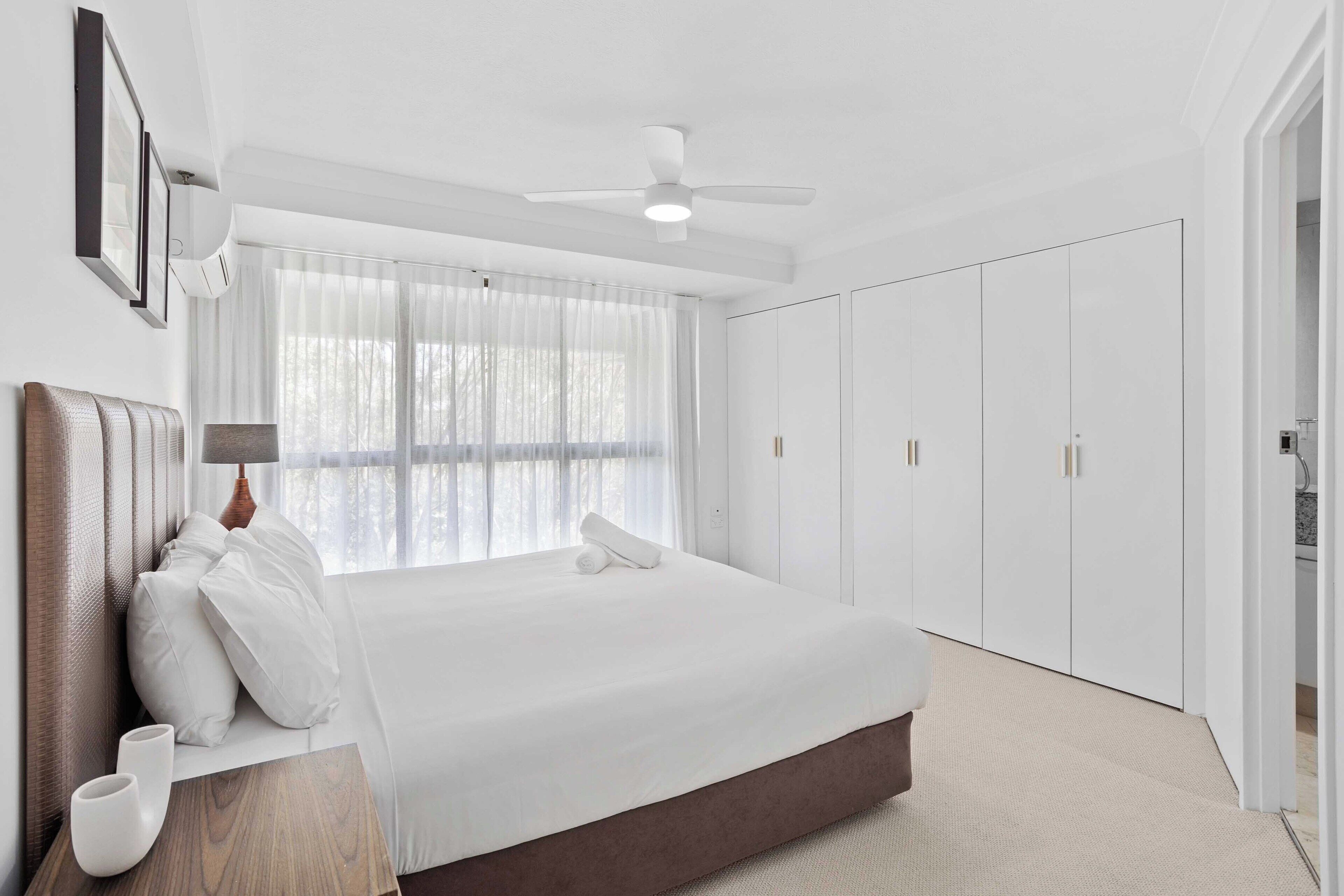Une chambre avec un grand lit, un ventilateur de plafond, une fenêtre avec des rideaux légers et une armoire.