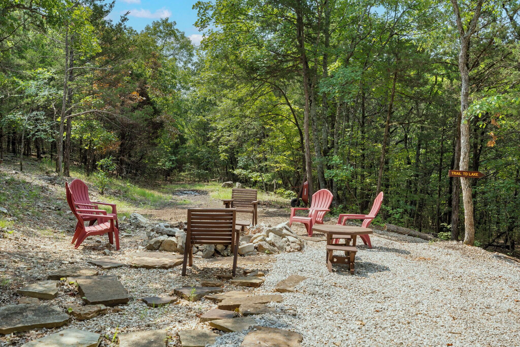 A forested area with a gravel path, red Adirondack chairs, a wooden bench, and a sign indicating a trail to a lake.