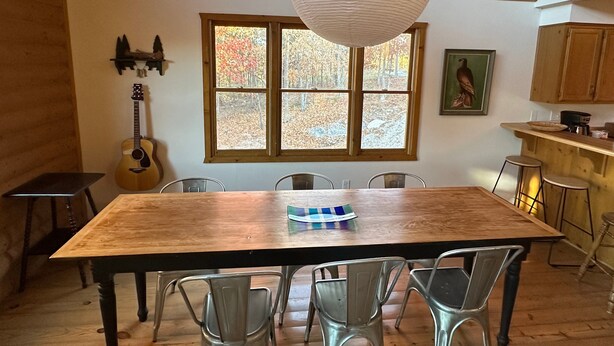 A wooden dining table with metal chairs, a guitar, and a large window with autumn foliage. - Private 4.5-Acre Retreat featuring Hot Tub-Firepit-Walk to Table Rock-Free Daily Attraction Tickets (Lampe)