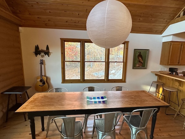 Una mesa de comedor de madera con sillas de metal, una guitarra y un ventanal con follaje otoñal.