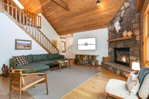A spacious living room with a green sofa, wooden coffee table, and a fireplace. A staircase with wooden railings leads to an upper level.