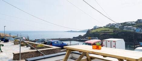 A coastal view with a wooden bench and picnic table, parked cars, and a trailer.