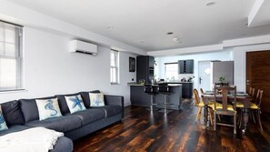 A modern living room with a dark wood floor, a grey sofa with decorative pillows, a dining area with wooden chairs, and a kitchen with dark cabinetry and a bar stool. - HipNautic, Mevagissey, sleeps 6 (Mevagissey)