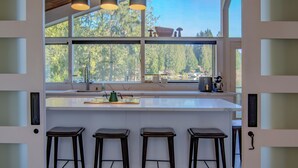Una cocina moderna con una isla grande, taburetes de bar y vistas a un bosque a través de amplios ventanales.