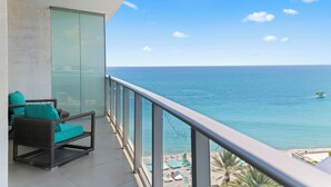 Un balcone con mobili in vimini e una porta in vetro, affacciato su un mare blu limpido e una costa lontana con ombrelloni da spiaggia.