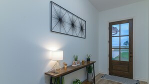 Ein Flur mit einem hölzernen Konsolentisch, einem an der Wand befestigten abstrakten Kunstwerk, einer Tür mit Blick ins Freie und einem kleinen Teppich auf dem Boden.