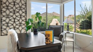 Une pièce avec une grande fenêtre, un bureau avec une lampe, une chaise et une plante en pot.