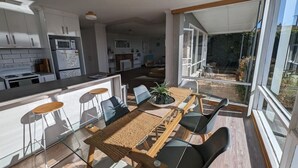 A modern kitchen with white cabinets, a microwave, and a refrigerator. There is a dining area with a table, chairs, and a plant. - Spacious Family Home in Devonport  (Devonport)