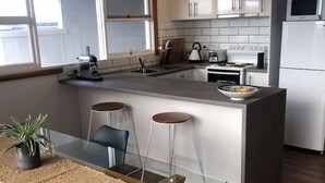 A modern kitchen with a central island, bar stools, a dining table with chairs, and a view of the outside through large windows. - Spacious Family Home in Devonport  (Devonport)