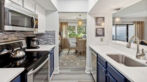A modern kitchen with white cabinets, a stainless steel microwave, and a coffee maker. There is a dining area with a table and chairs, and a view of the outdoors through a window. - 266 Turnberry Village @ Palmetto Dunes | Golf View | Walk to Beach | Pool Access (Hilton Head Island)