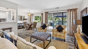 A modern living room with a sofa, ottoman, and striped armchairs, a dining area with a table and chairs, and a kitchen in the background. - 266 Turnberry Village @ Palmetto Dunes | Golf View | Walk to Beach | Pool Access (Hilton Head Island)