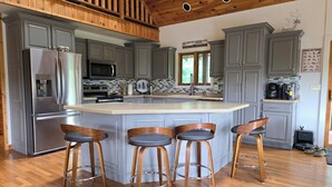 Una cucina moderna con mobili grigi, un frigorifero in acciaio inossidabile, un forno a microonde e una macchina da caffè. Quattro sgabelli da bar in legno con gambe in metallo.