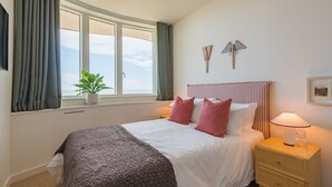 A neatly made bed with a white and gray blanket, a bedside lamp, and a window with curtains.