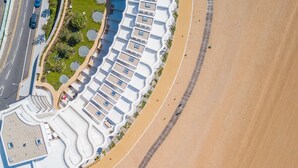 An aerial view of a coastal hotel with a curved building layout, a sandy beach, and a road with vehicles.