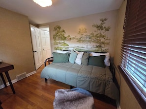 A bedroom with a bed, a chair, a small table, a window with blinds, and a door.