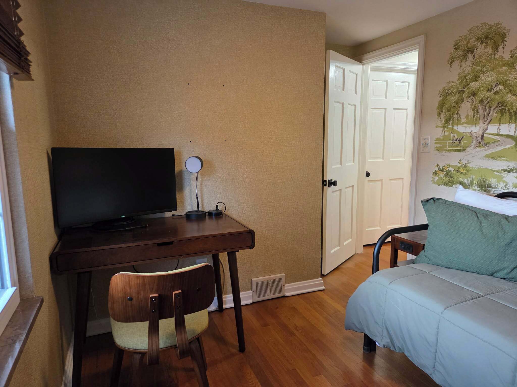 A bedroom with a wooden desk, a chair, a bed, and a door leading to another room.
