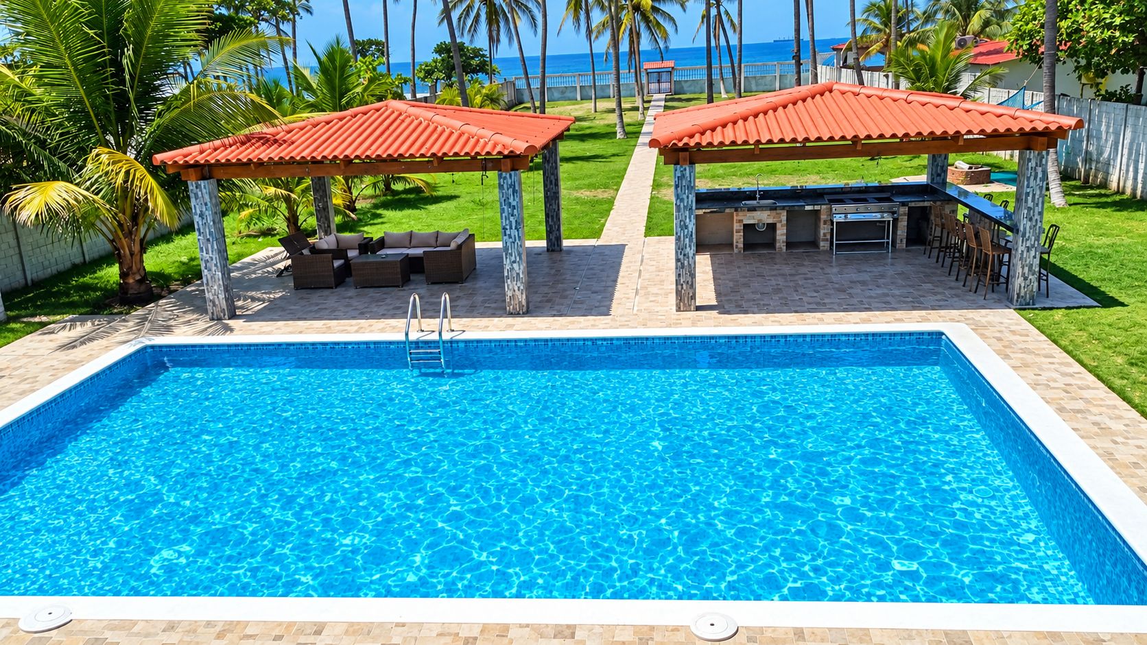 Ein rechteckiger Pool mit einer gefliesten Liegefläche, zwei überdachten Sitzbereichen mit roten Dächern und Blick aufs Meer.