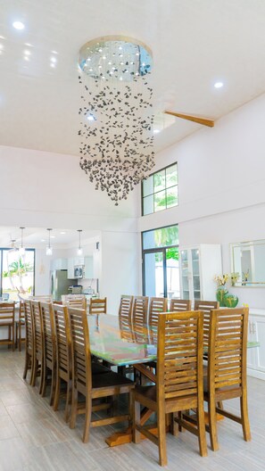 Un vaste espace dédié à la salle à manger, avec une grande table ronde entourée de chaises en bois, un lustre moderne et une vue sur la cuisine et l’extérieur grâce à de grandes fenêtres.