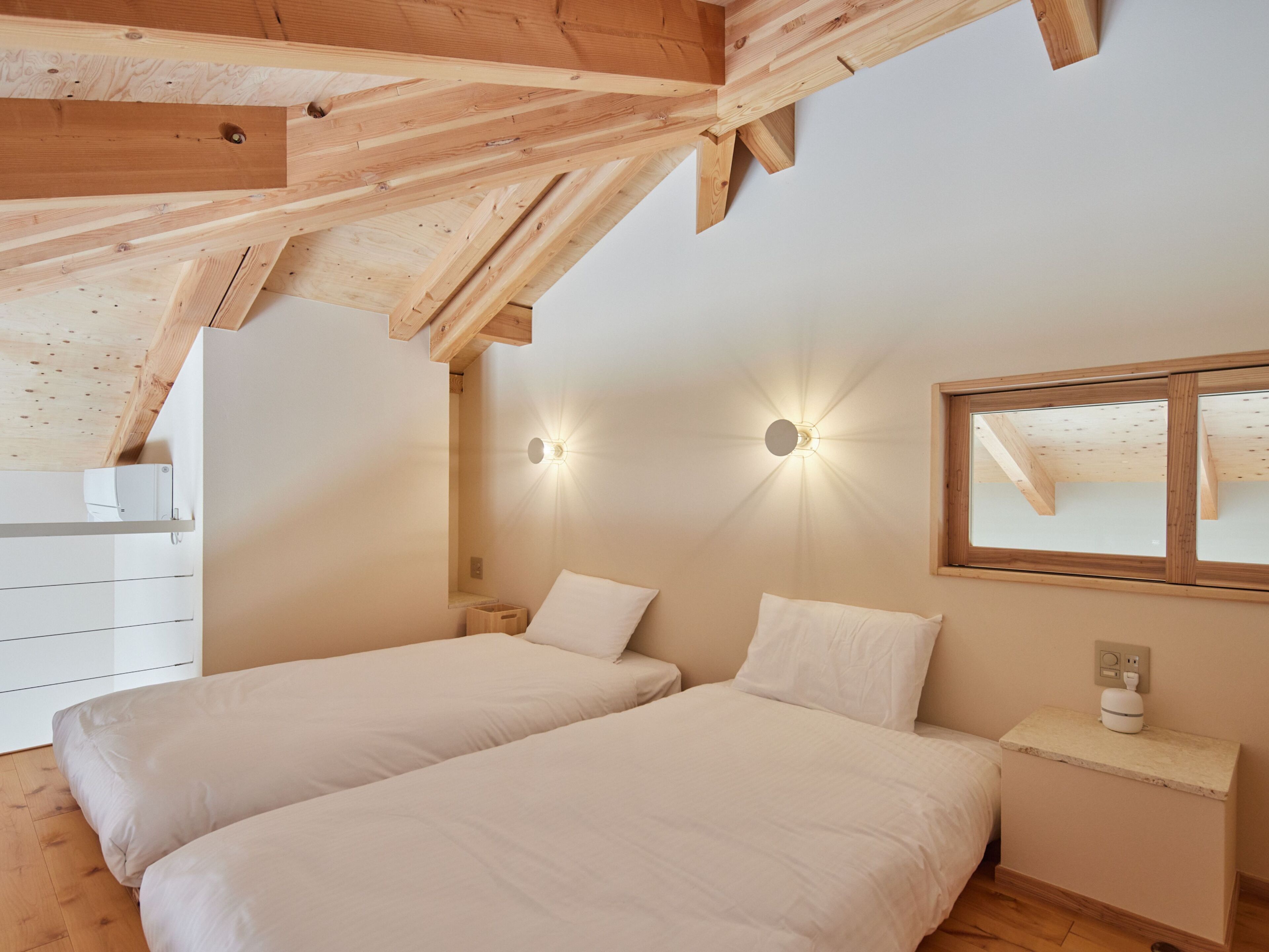 Une chambre avec deux lits, des poutres apparentes, une lampe fixée au mur et une fenêtre.