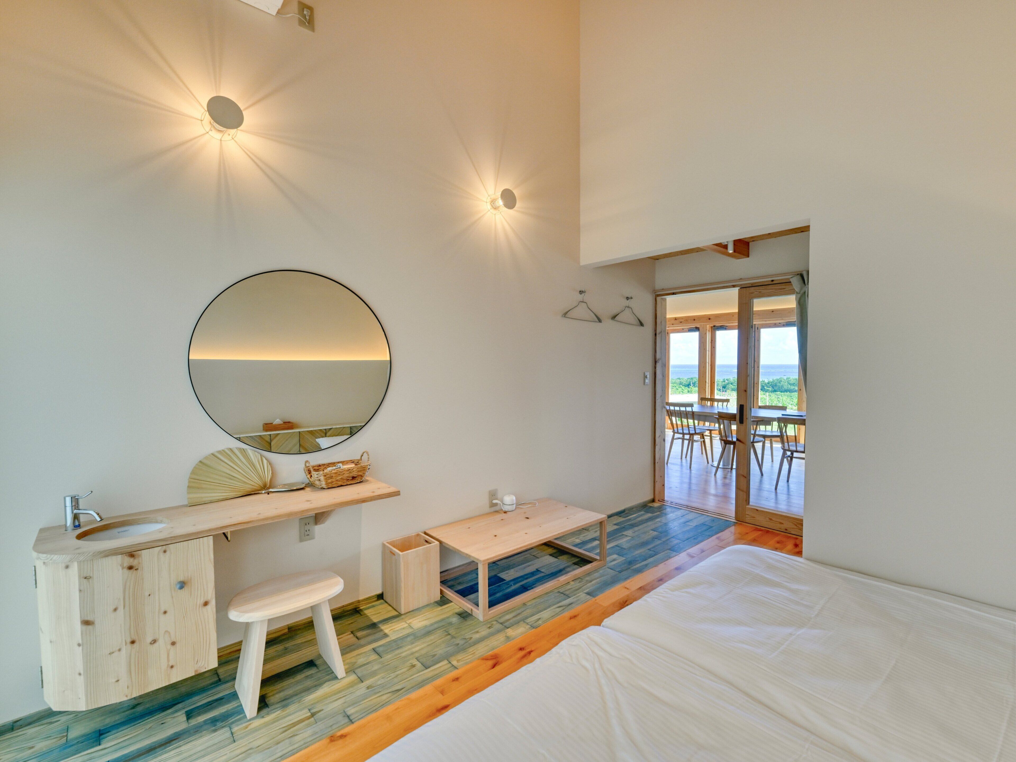A bedroom with a wooden bed, a round mirror, a small desk with a chair, a wooden bench, and a view of the outdoors through a glass door.