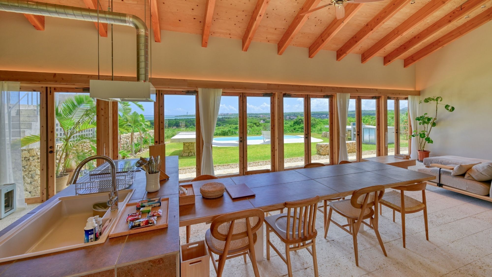 Een ruime keuken met een groot eiland, een eethoek met stoelen, en uitzicht op een zwembad en groen.