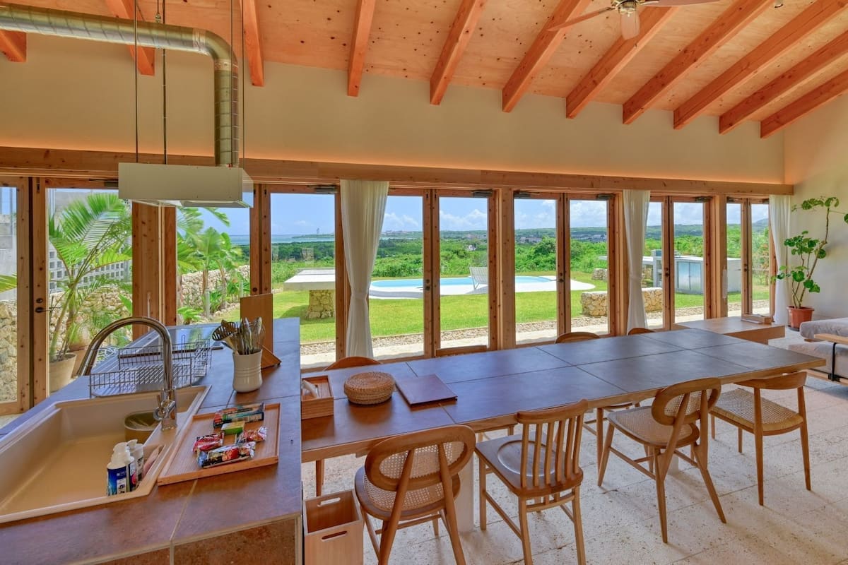 A spacious kitchen with a large island, a dining area with chairs, and a view of a pool and greenery.