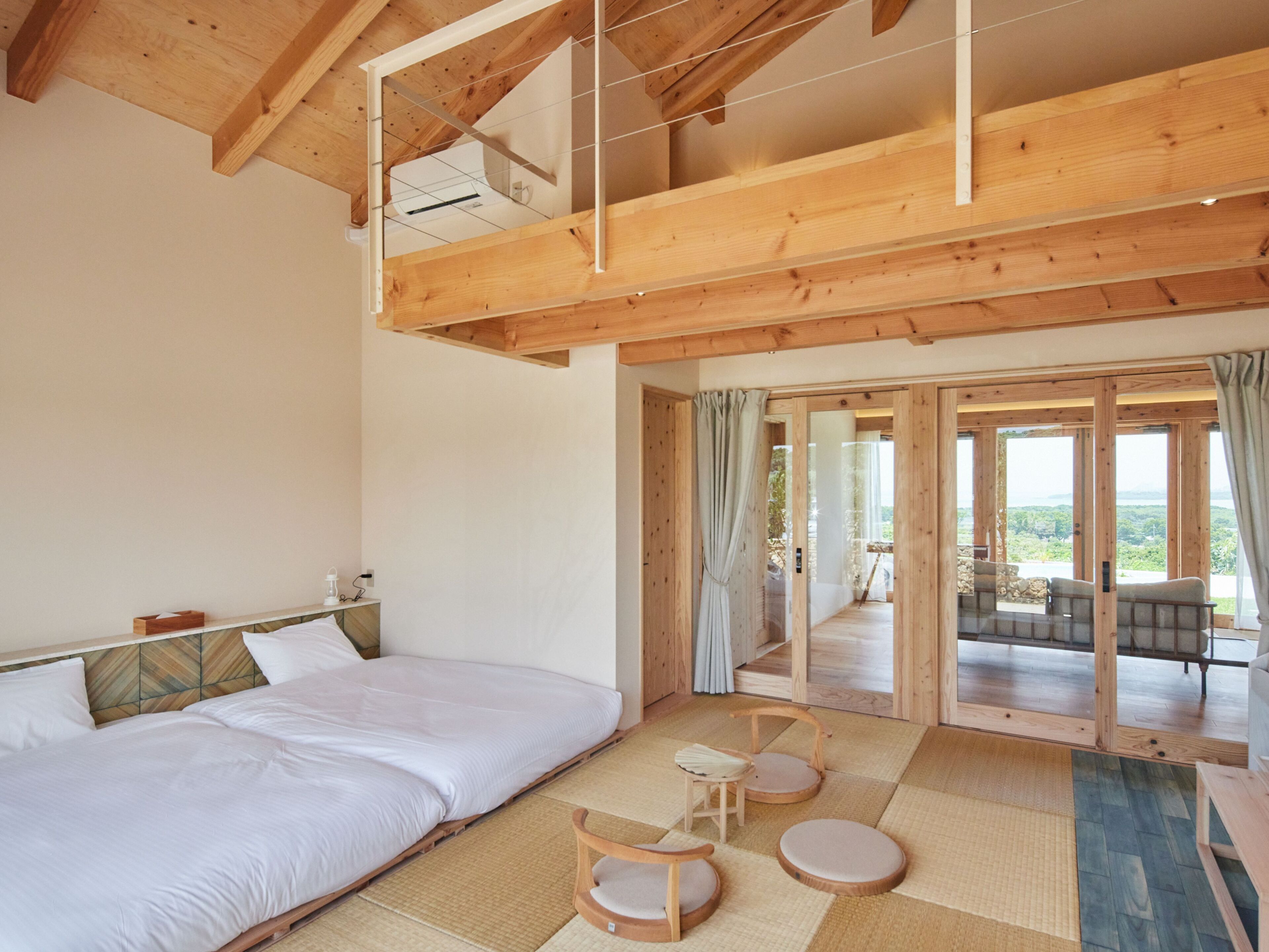 A spacious room with a large bed, a small table, and a chair. There is a view of the outdoors through a glass door.