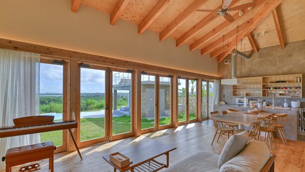 A spacious living area with a piano, a sofa, a dining table, and a kitchen area with wooden cabinets and a stone wall. - Ishigaki hills (Ishigaki)