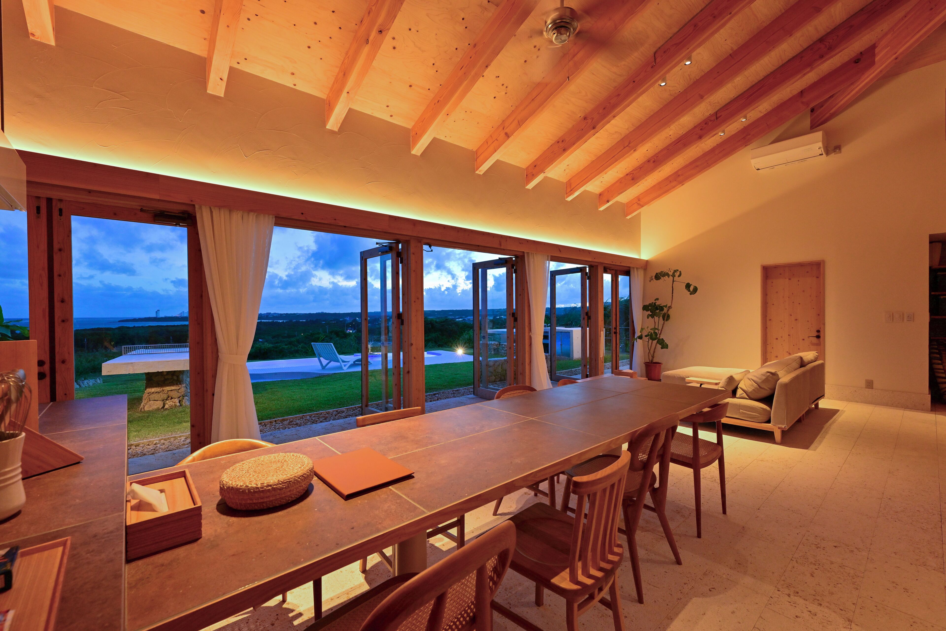 A spacious dining area with a long wooden table, multiple chairs, and large windows offering a view of the outdoors.