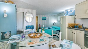 Une pièce comprenant une table à manger en verre, un lit avec une literie bleue, un coin cuisine et une salle de bain avec des meubles blancs.