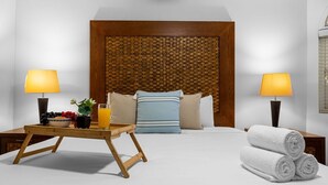 Une chambre d’hôtel avec un grand lit, deux lampes de chevet, une tête de lit en bois, un plateau avec des fruits et un verre de jus, et des serviettes roulées sur le côté.