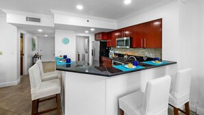 Une cuisine moderne avec des meubles de rangement en bois foncé, un four à micro-ondes en acier inoxydable et un plan de travail noir. On y trouve deux chaises blanches et un set de table bleu sur le plan de travail.