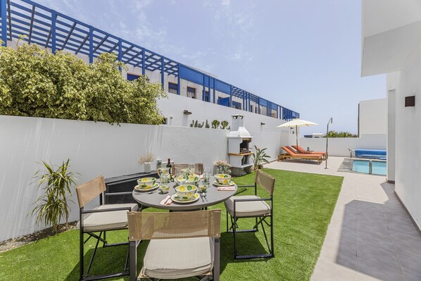 A patio with a round table set for dining, surrounded by wicker chairs, and a pool area with lounge chairs. - Villa Cartali Playa Blanca (Playa Blanca)