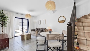 Ein modernes Wohnzimmer mit einem runden Esstisch, zwei Stühlen, einem Sofa und einem Couchtisch. Eine Treppe mit Büchern an der Seite und ein großer Spiegel an der Wand sind ebenfalls vorhanden.