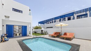 Ein modernes Außenpool-Areal mit einem rechteckigen Pool, blauen Rollläden und zwei Liegestühlen mit Sonnenschirm.