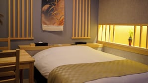 Ein Hotelzimmer mit Bett, Schreibtisch, Stuhl, Nachttisch, Wandlampe und einem Fenster mit Pflanze.