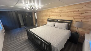 A bedroom with a wooden headboard, a bed with a grey bedspread, a nightstand with a lamp, and a wooden floor. - Gibson Getaway- Relax and Unwind (Wagoner)