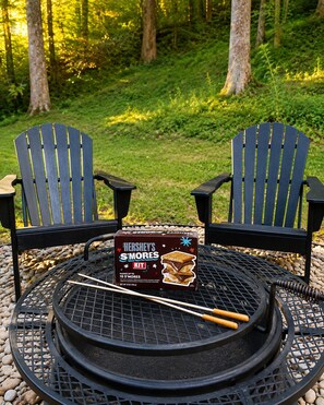 Two Adirondack chairs, a Hershey's S'mores kit, and a round metal grill on a table surrounded by pebbles.