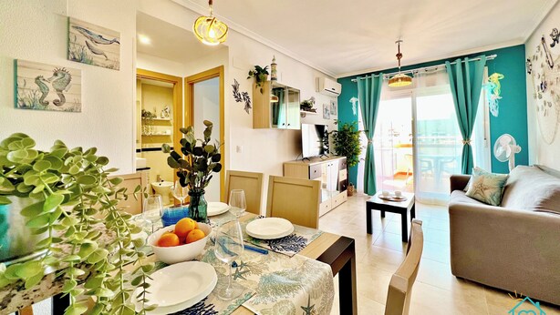 Ein modernes Wohnzimmer mit einer Couch, einem Esstisch gedeckt mit Tellern und Gläsern, einer Vase mit Blumen und Blick ins Freie durch große Fenster.