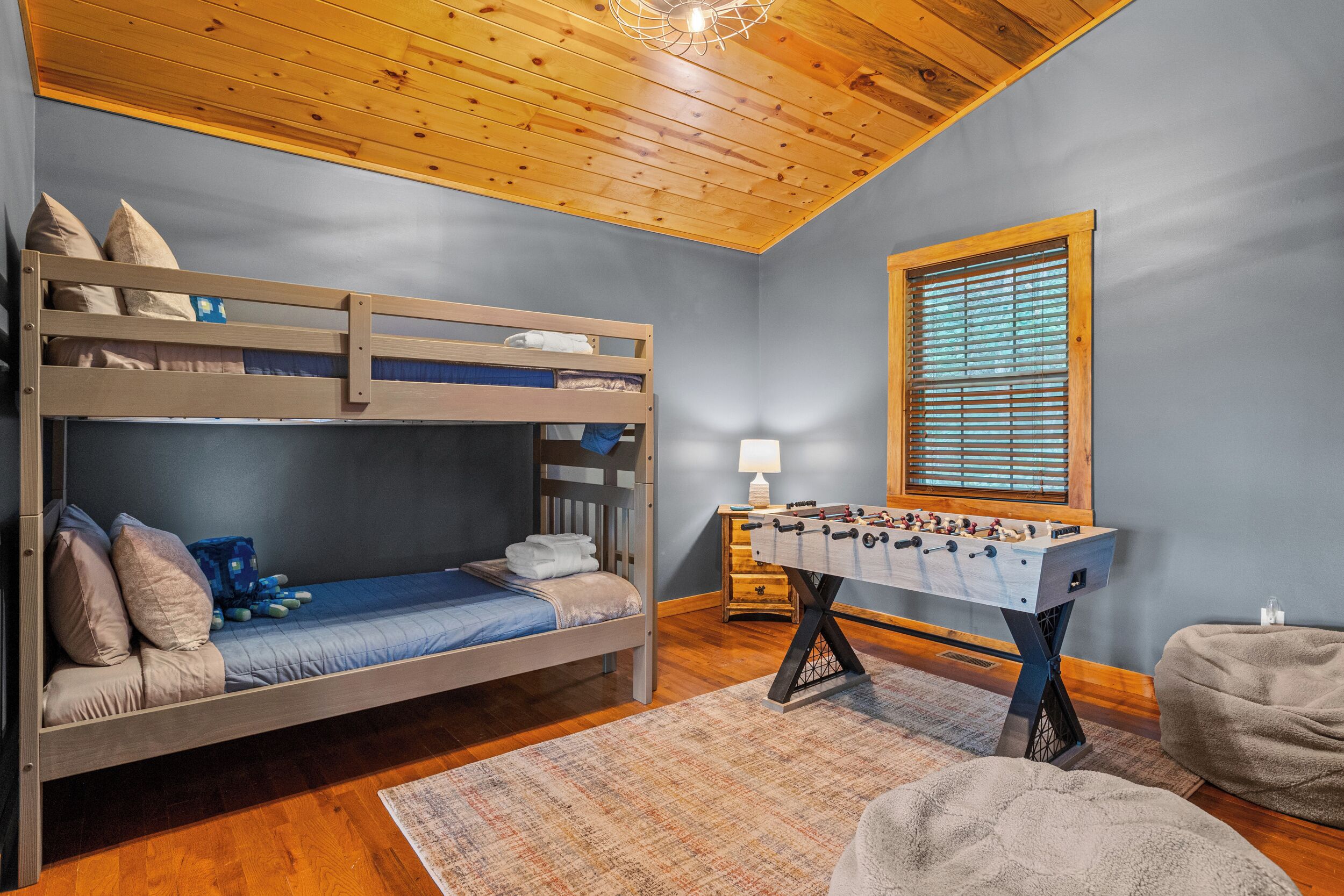 Un lit superposé avec des oreillers et une couverture bleue, une table de baby-foot, une lampe et un fauteuil en forme de poire dans une pièce au plancher en bois et au plafond en pente.